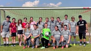 Girls' youth soccer team in gray uniforms posing with two coaches in front of a goal on a sunny field.