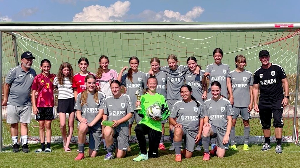 Girls' youth soccer team in gray uniforms posing with two coaches in front of a goal on a sunny field.
