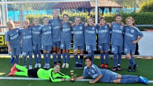Youth soccer team in blue uniforms posing for a group photo on the field; two goalkeepers lie in front with a trophy between them.