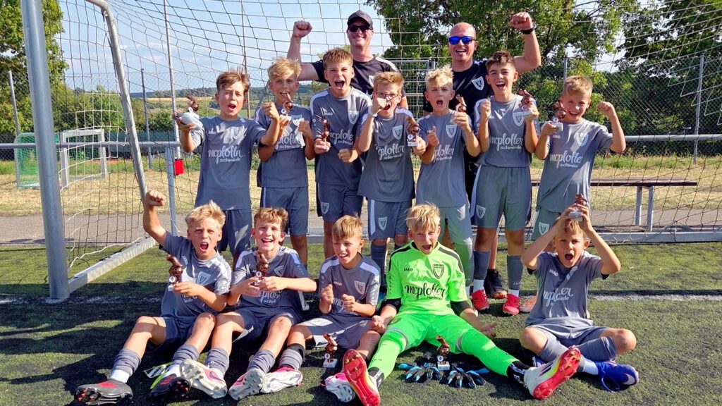 Youth soccer team in gray uniforms celebrate victory with trophies, flanked by two cheering coaches near the goal.
