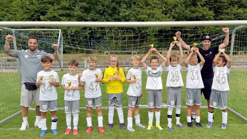 Youth soccer team posing with trophies in front of a goal after a match celebration on a grassy field, with two coaches at each end raising their arms in triumph.