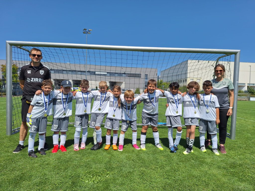 Group of young soccer players wearing white uniforms with medals, posing in front of a goal on a sunny field with coaches at each end.