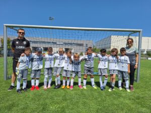 Group of young soccer players wearing white uniforms with medals, posing in front of a goal on a sunny field with coaches at each end.