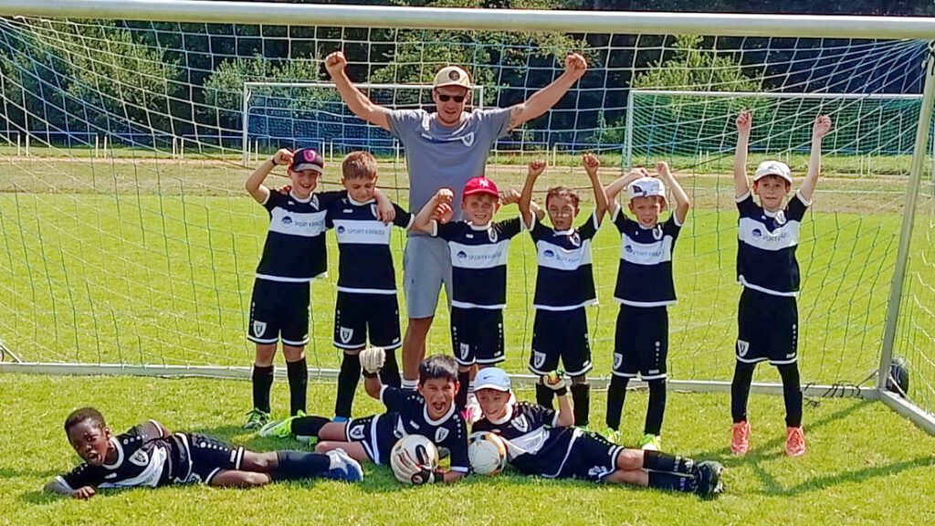 Youth soccer team posing in front of a goal with a coach, arms raised in celebration on a sunny field.