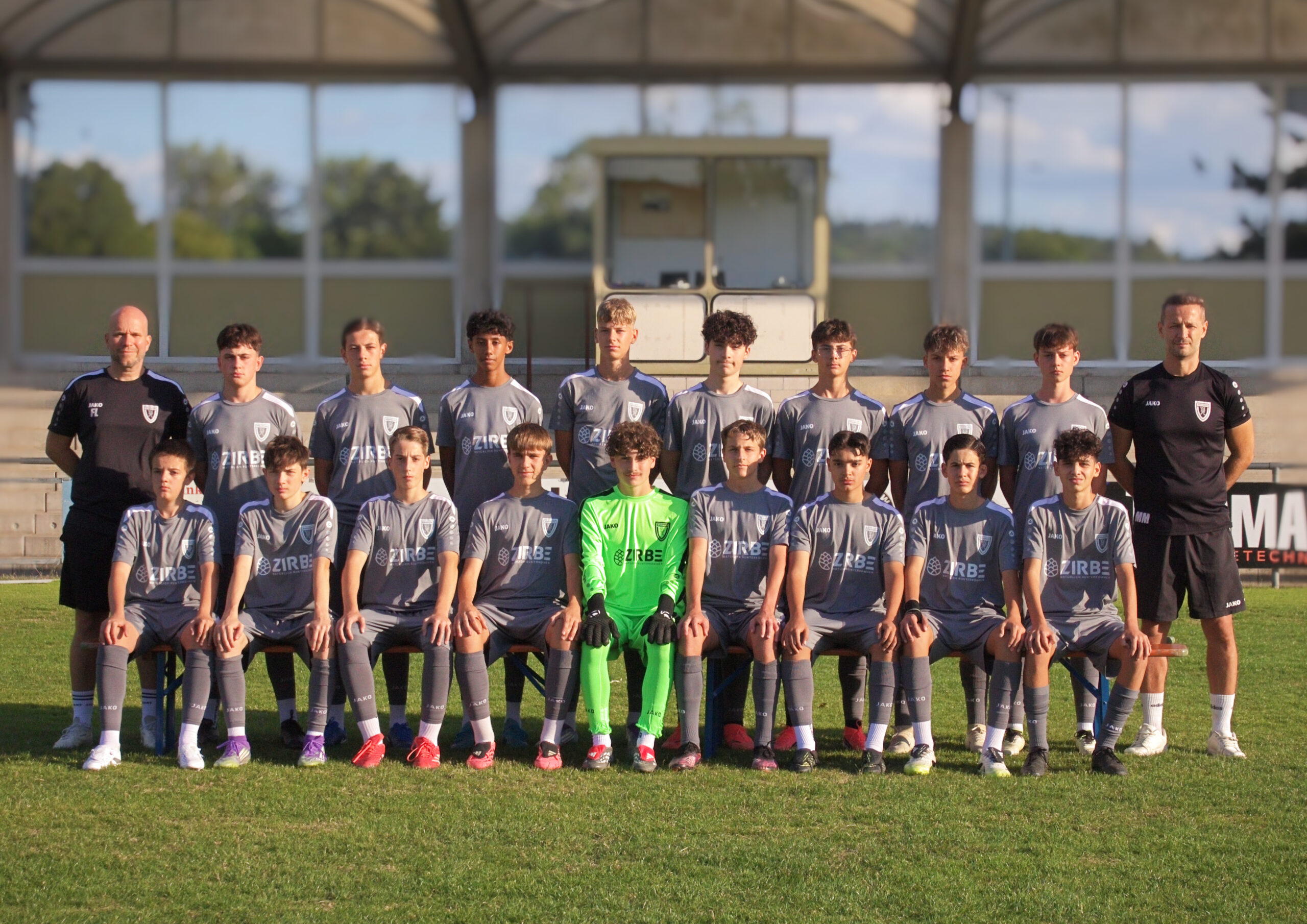Youth soccer team in gray uniforms posing for a group photo on the field, with two coaches at the ends and a neon-green goalkeeper in front center.