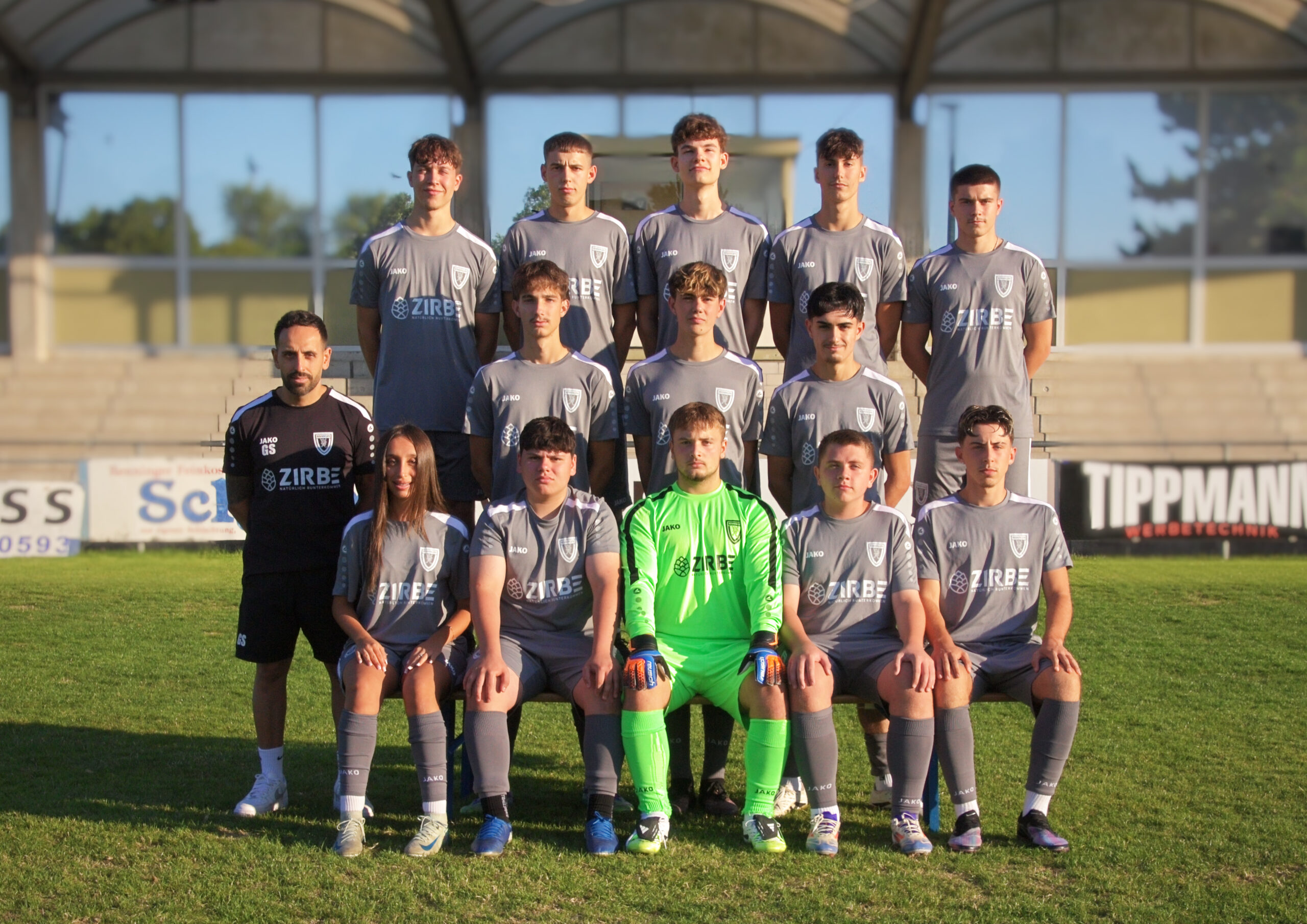 Soccer team posing for a group photo on a grass field, players in gray jerseys with a neon-green keeper, stadium behind them.