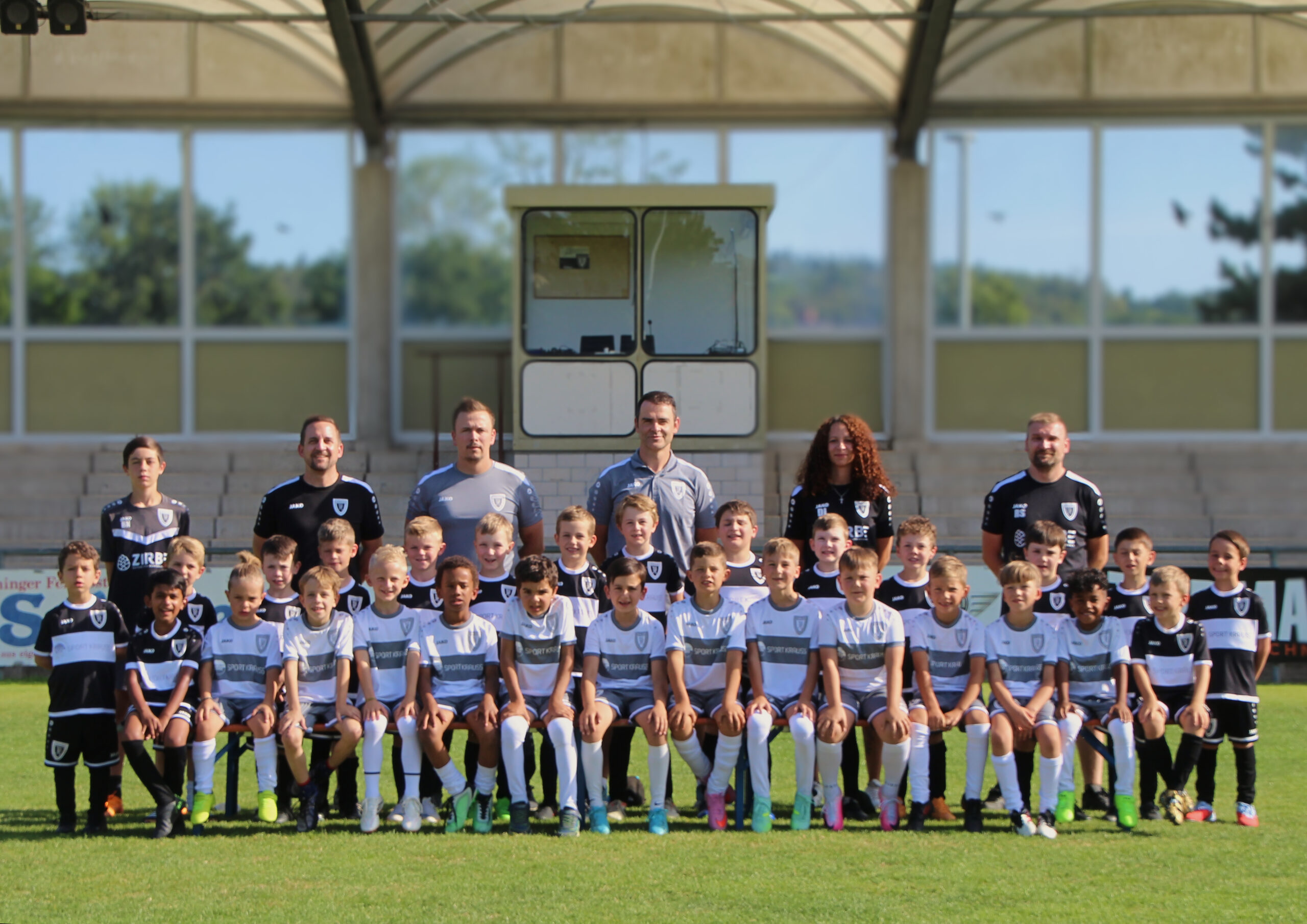 Group photo of a soccer team and children posing on a field with stadium stands behind it.