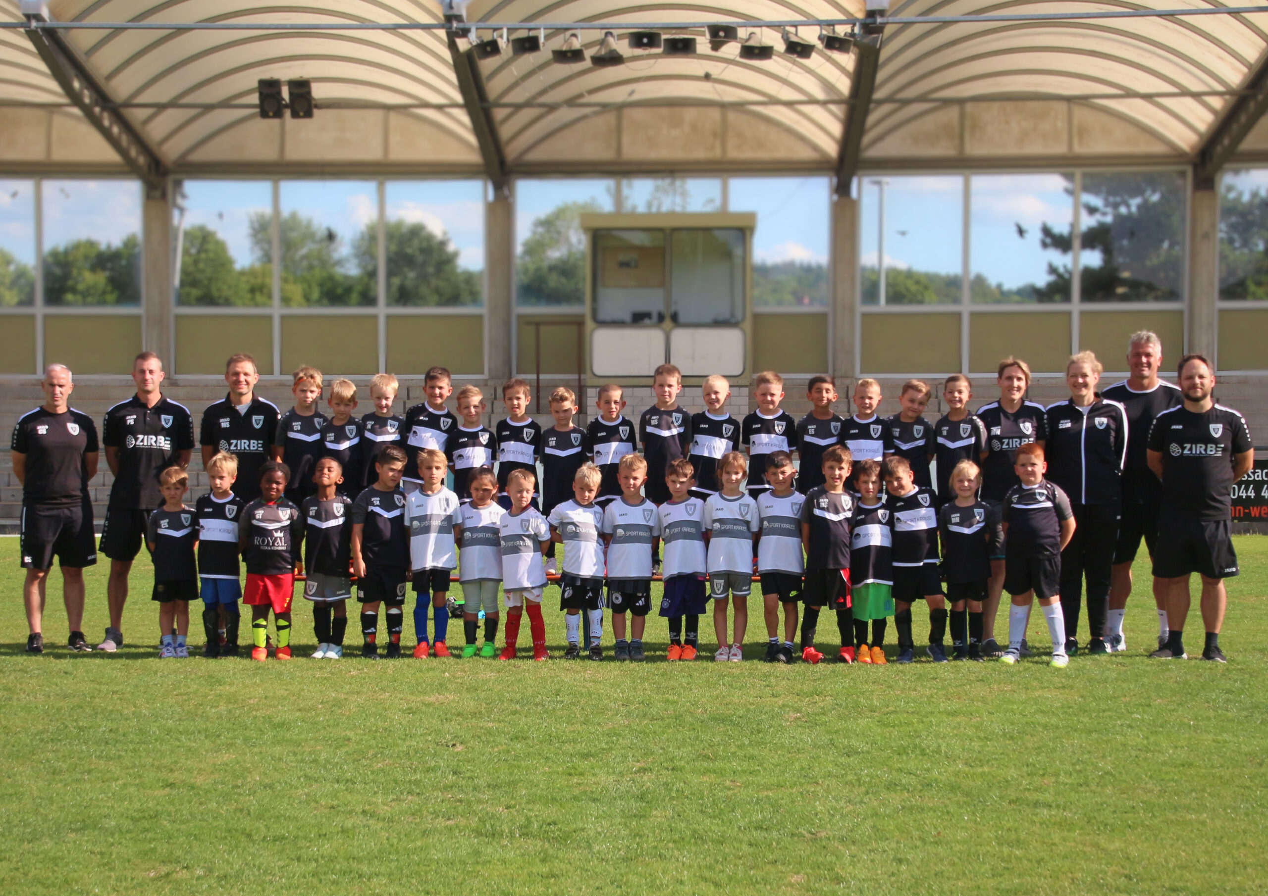 Group of young soccer players and coaches in black-and-white uniforms posing for a team photo on a sunny field under a curved glass-roofed structure.