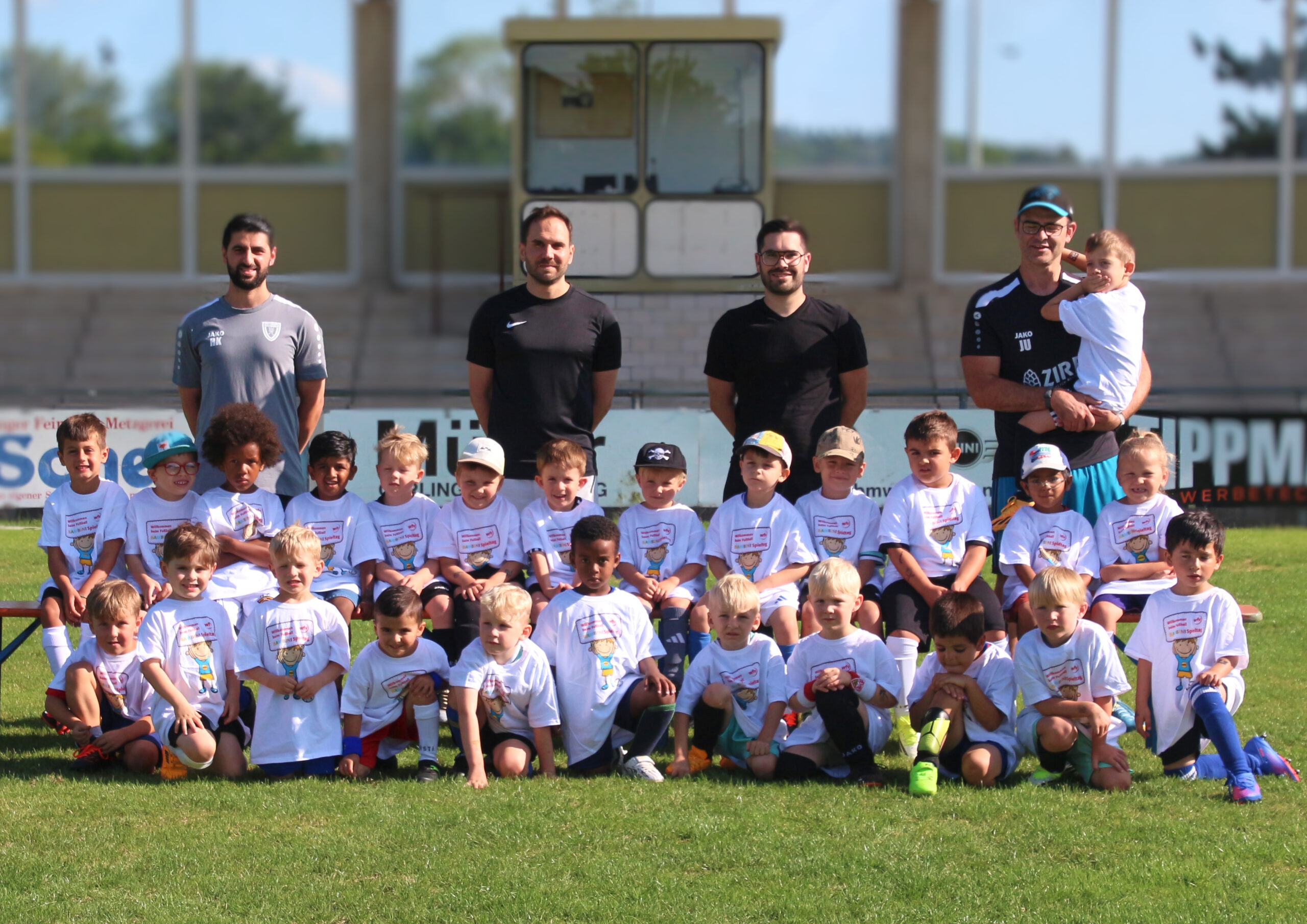 Group of young kids in white shirts posing for a team photo on a sunny sports field, with several adults standing behind them.