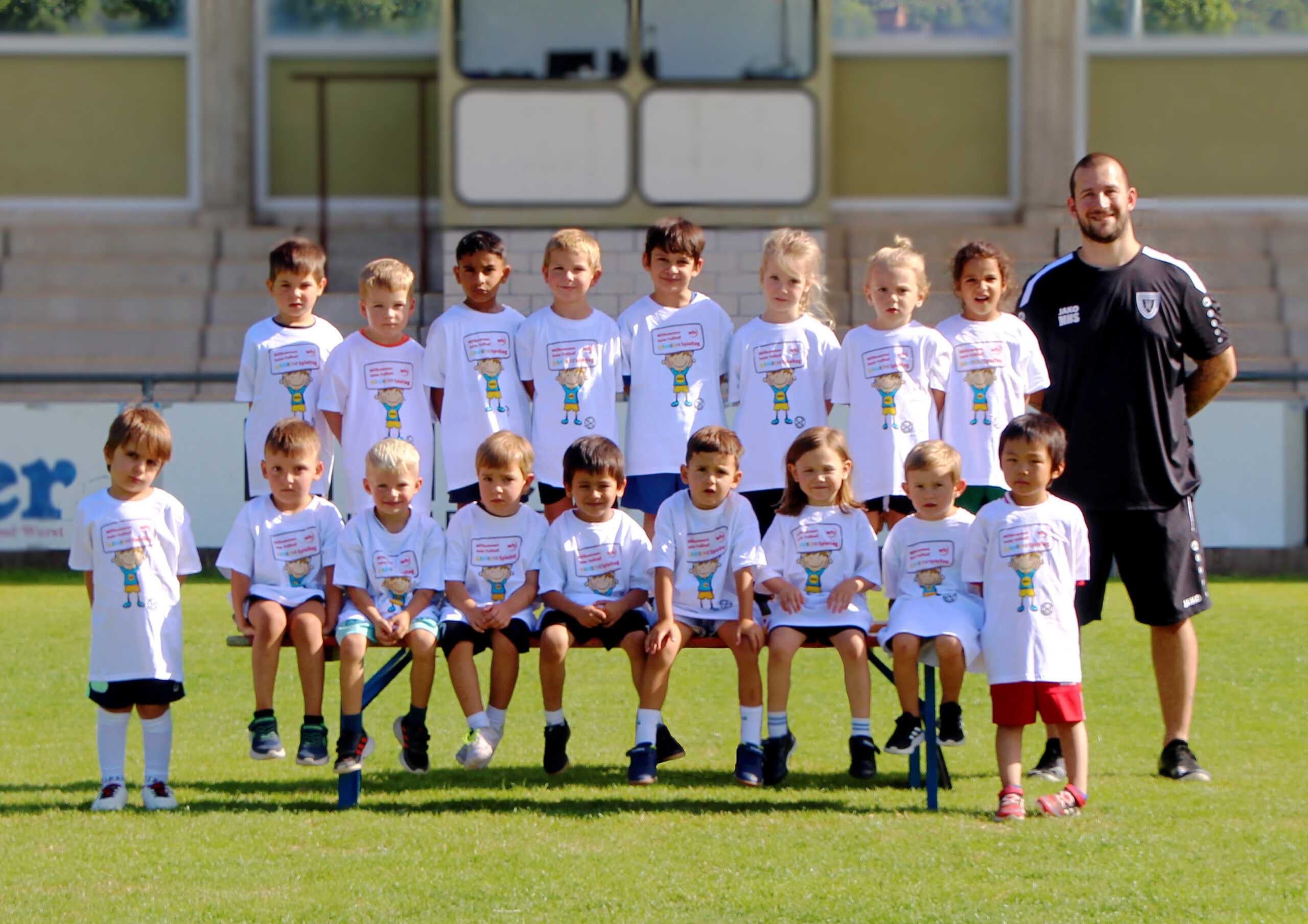 Group of children in white shirts posing for a team photo on a grassy field, with a coach on the right.