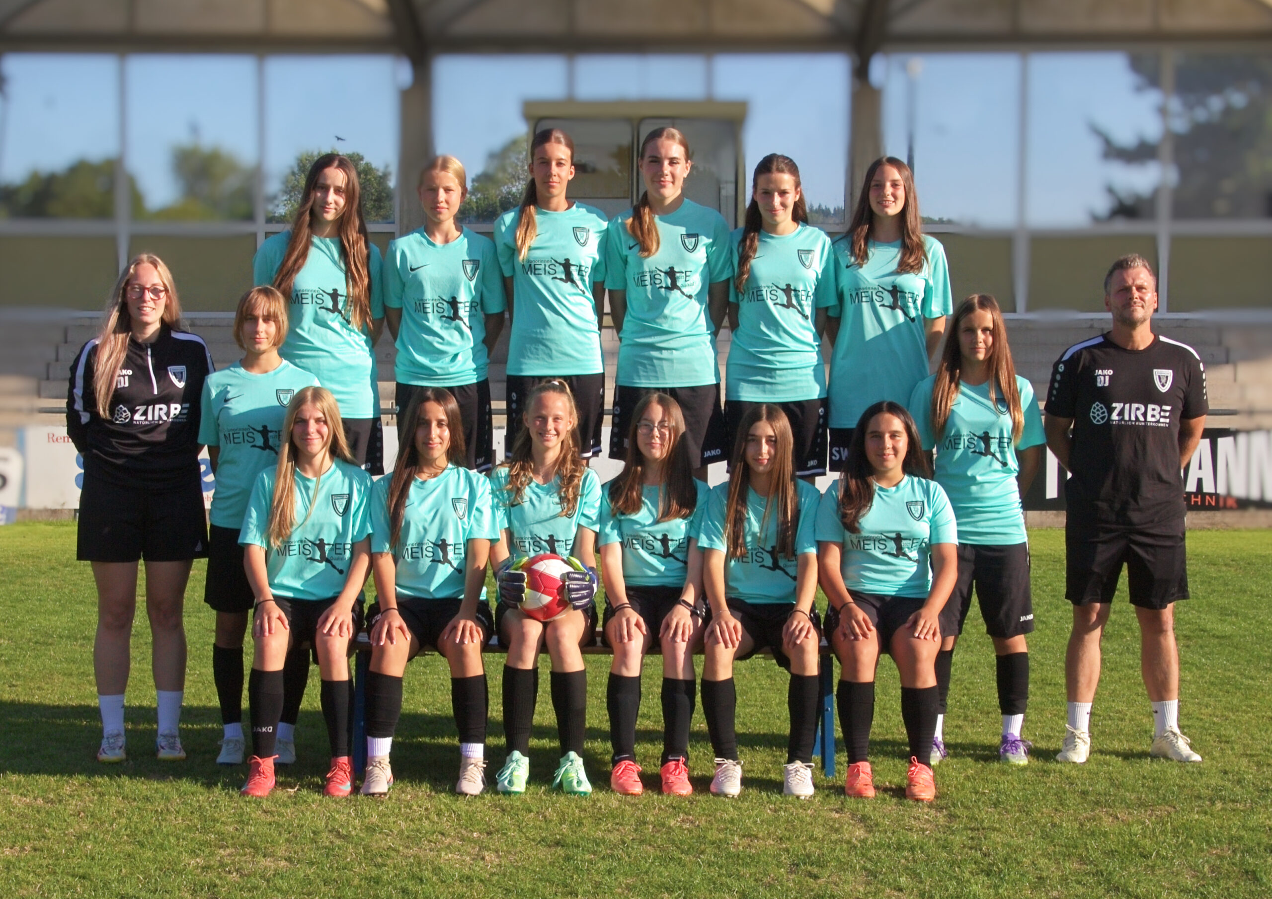 Group of young women soccer players in teal jerseys posing on the grassy field with two coaches on either end.