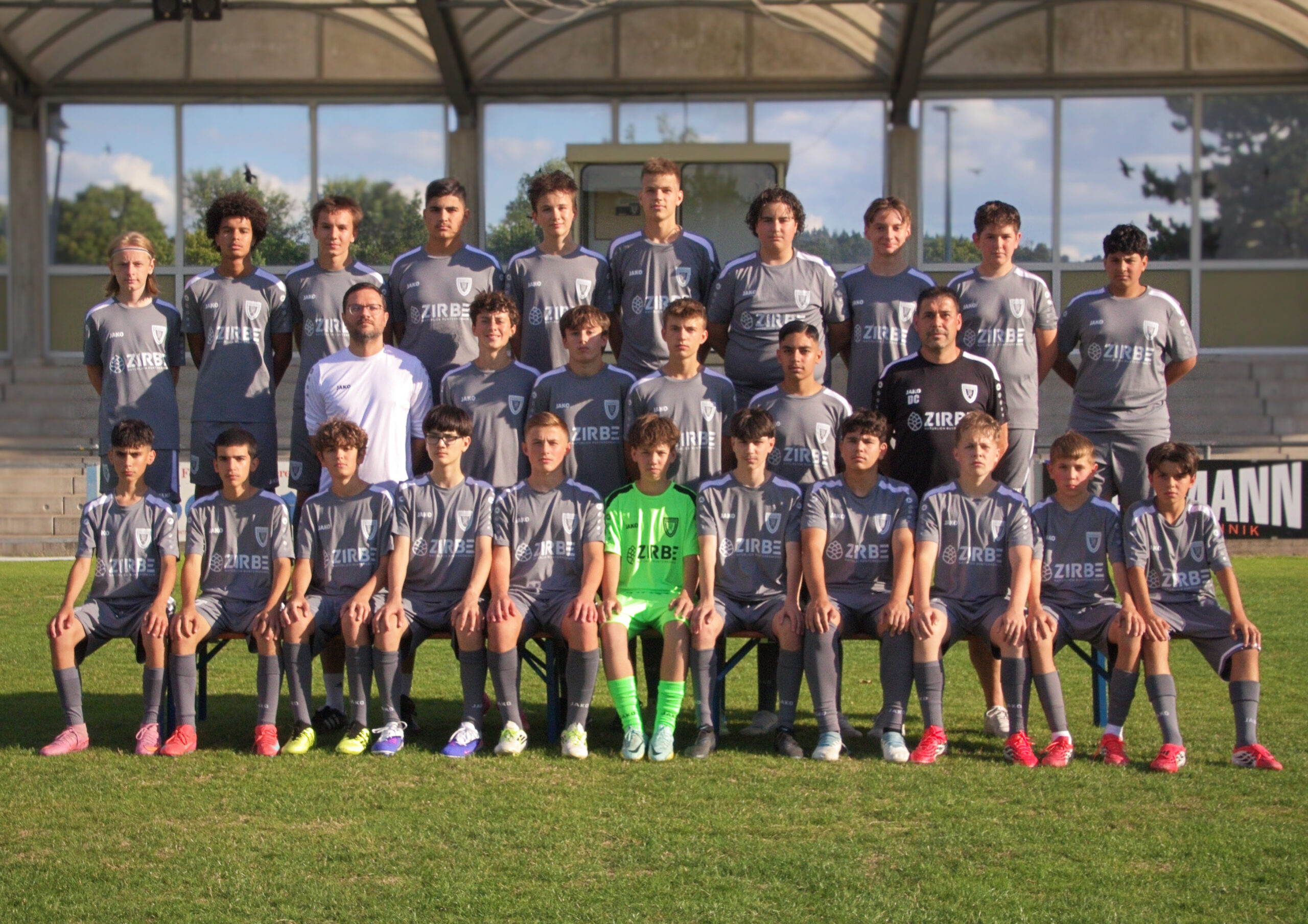 Youth soccer team posing for a group photo on bleachers in matching gray kits with sponsor logos and a bright green goalie in front row.