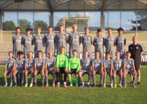 Youth soccer team posing for a group photo on the field in gray uniforms with a coach at the right.