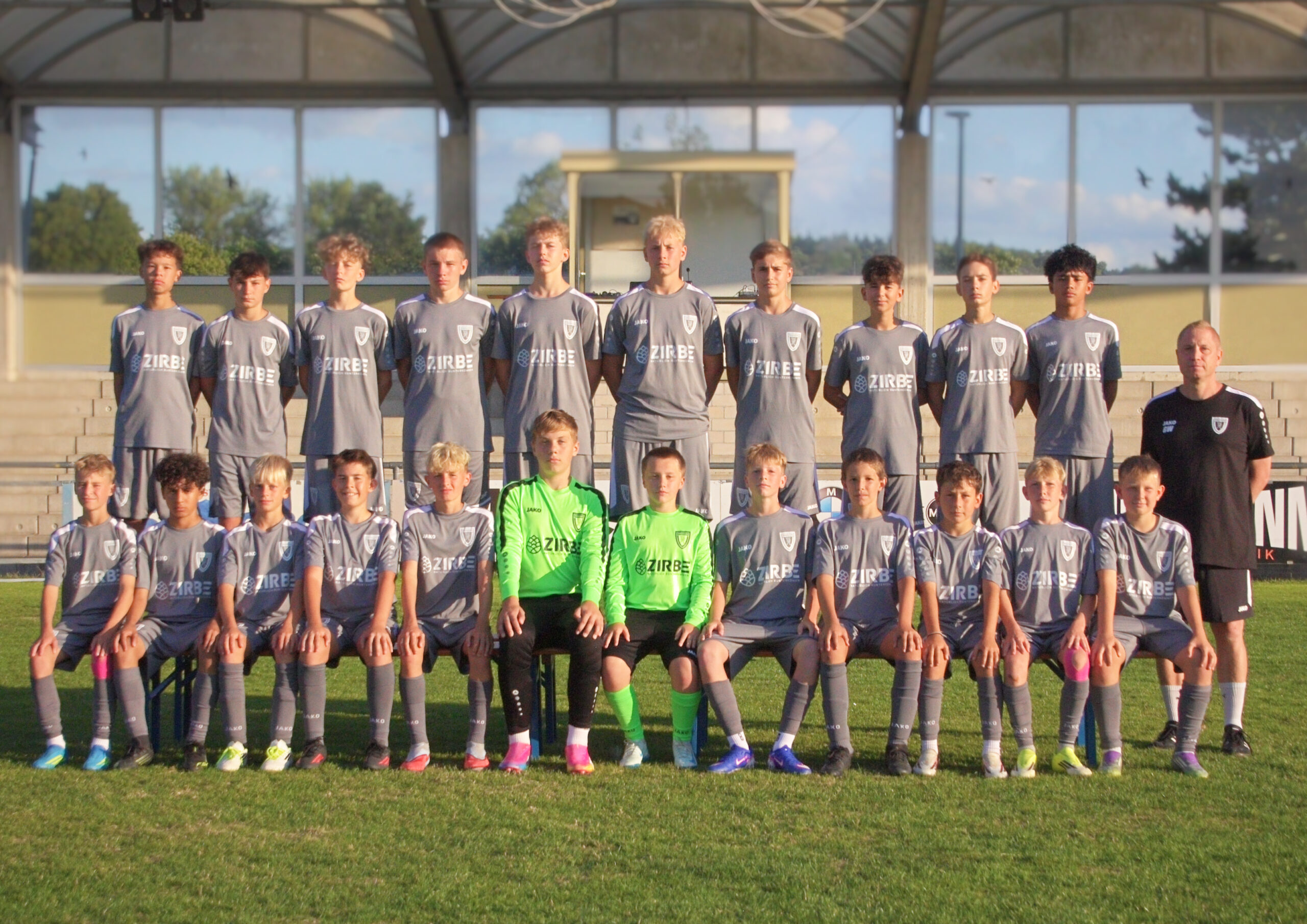 Youth soccer team posing for a group photo on the field in gray uniforms with a coach at the right.