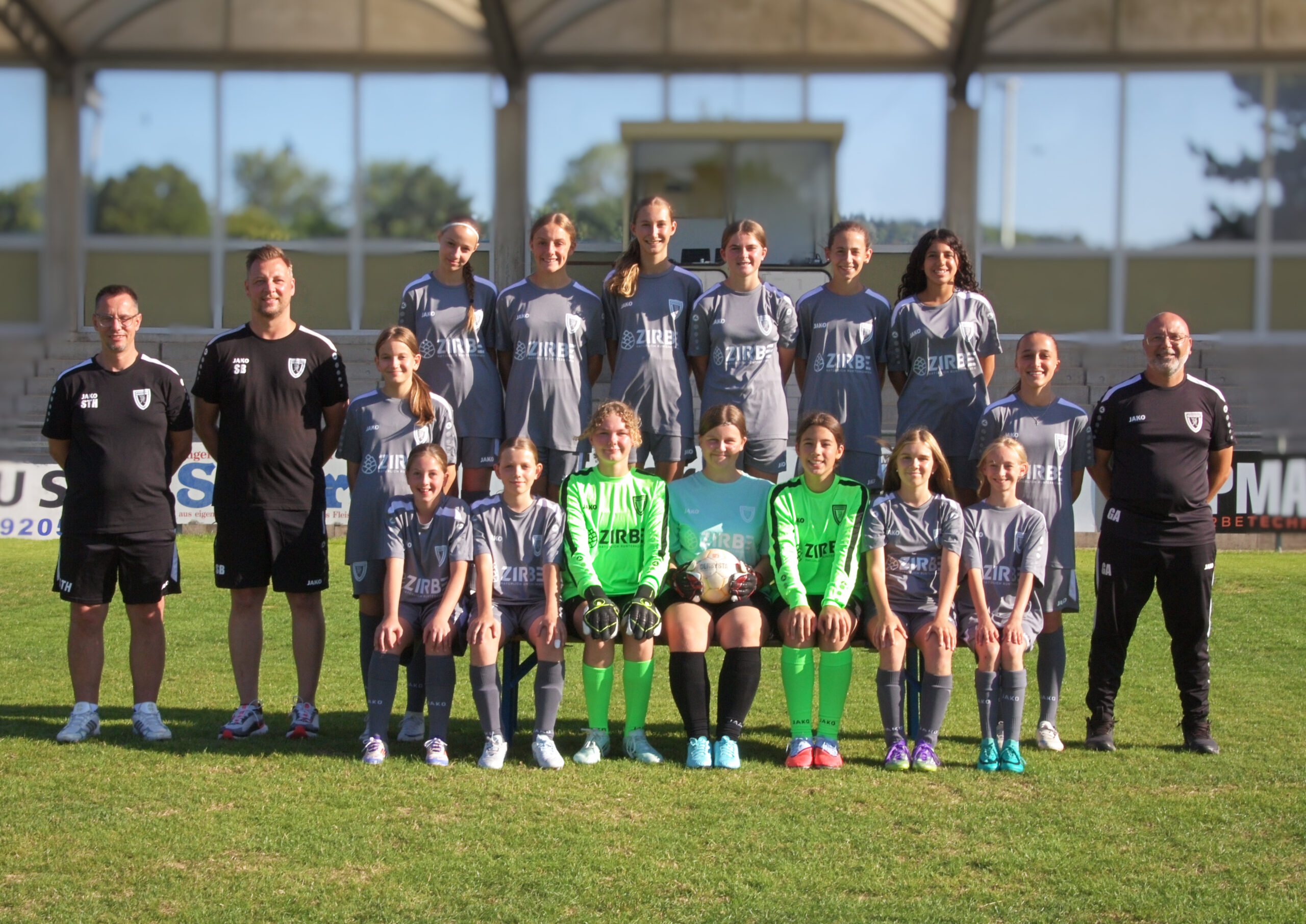 Group of girls and coaches in gray soccer uniforms posing for a team photo on a sunny field, front row seated, goalkeepers in neon green jerseys.