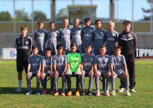 Group of young soccer players in gray uniforms posing for a team photo on a grassy field; two adults in dark jackets stand at sides.