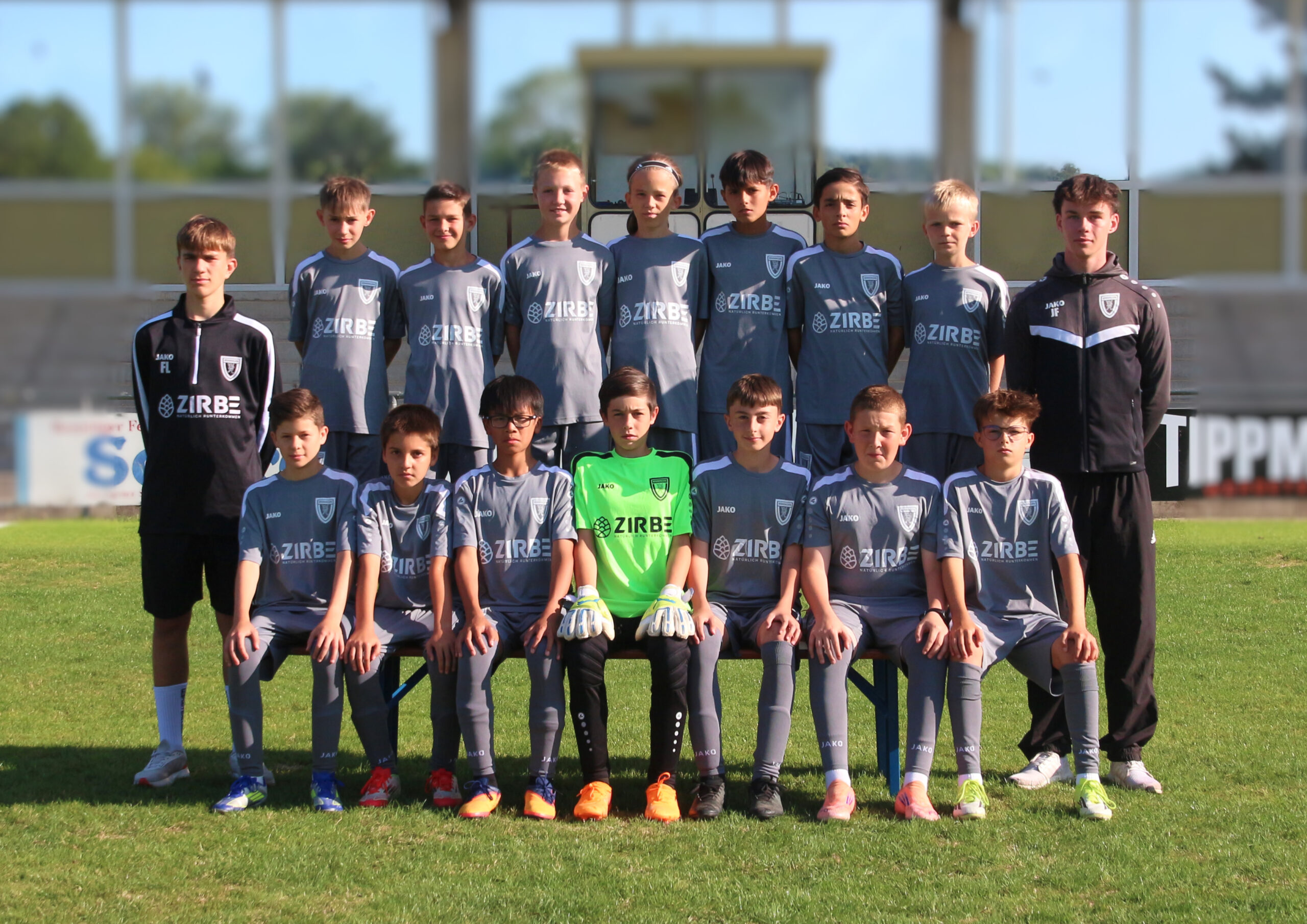 Group of young soccer players in gray uniforms posing for a team photo on a grassy field; two adults in dark jackets stand at sides.