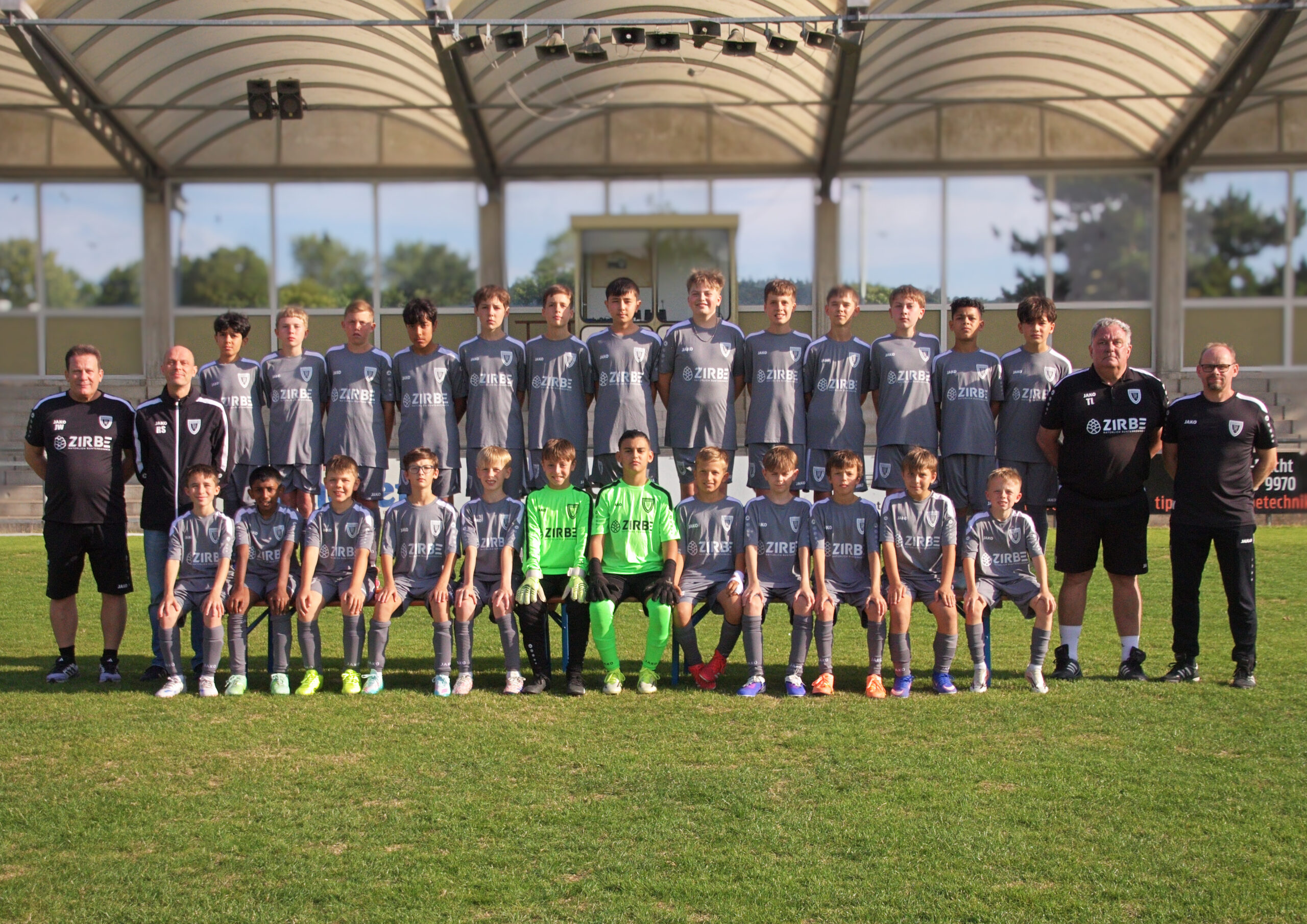 Youth soccer team and coaches posing for a group photo on a sunny day, wearing gray jerseys and standing on a grassy field with stadium seating behind them.