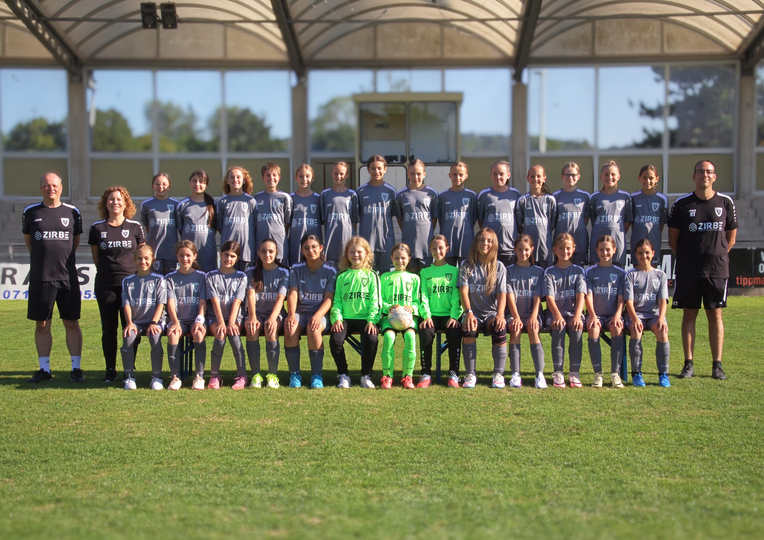 Group photo of a girls' soccer team in gray jerseys with neon-green goalkeepers, flanked by two coaches on a sunny field.
