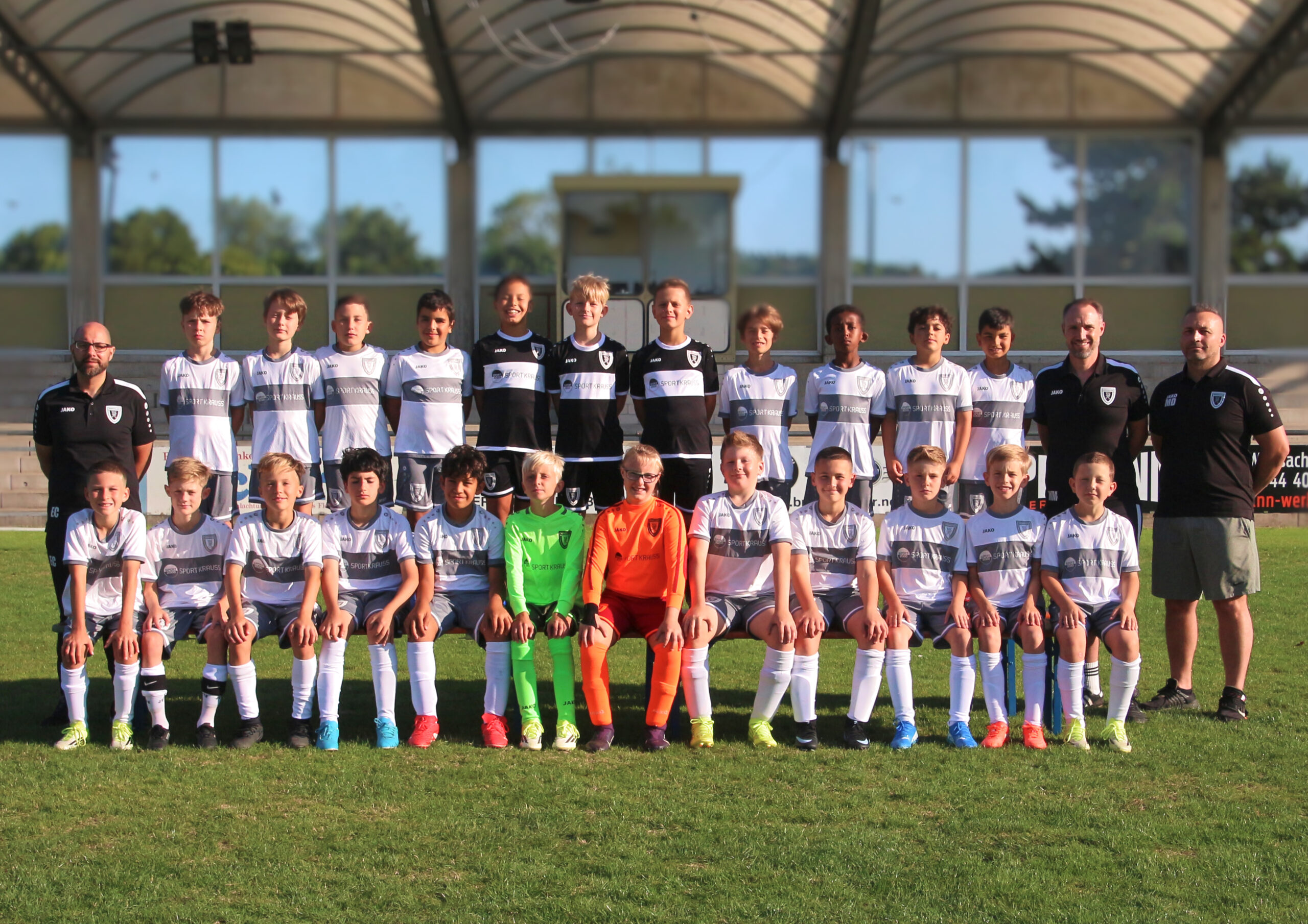 Group of youth soccer players and coaches posing on the field in matching white uniforms.