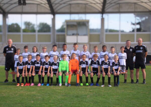 Youth soccer team posing for a group photo on a grass field with two coaches at the ends.