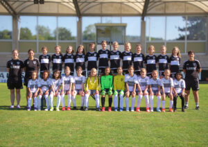Group of young female soccer players in black-and-white uniforms posing with two coaches on a sunny field stadium backdrop.