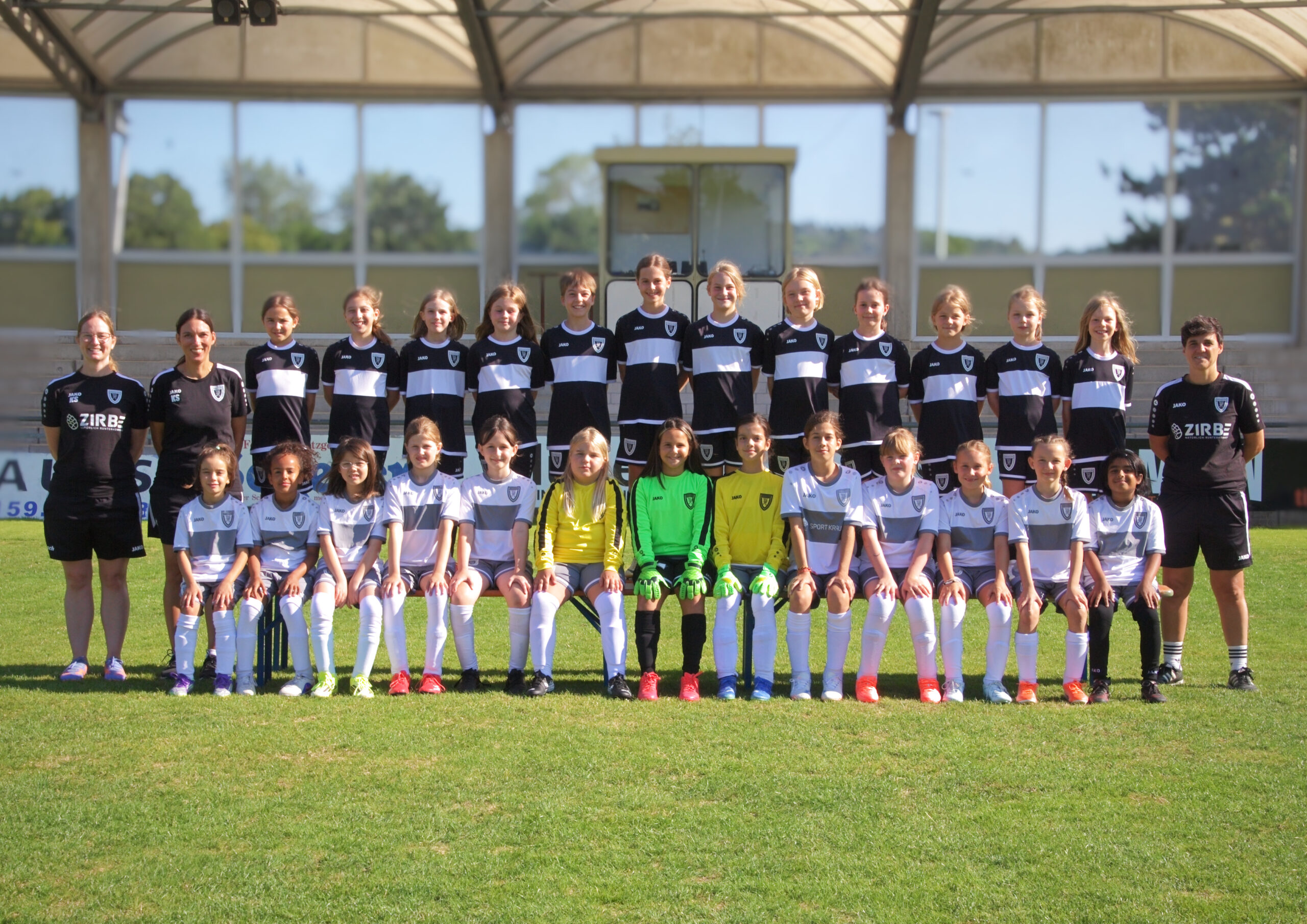 Group of young female soccer players in black-and-white uniforms posing with two coaches on a sunny field stadium backdrop.
