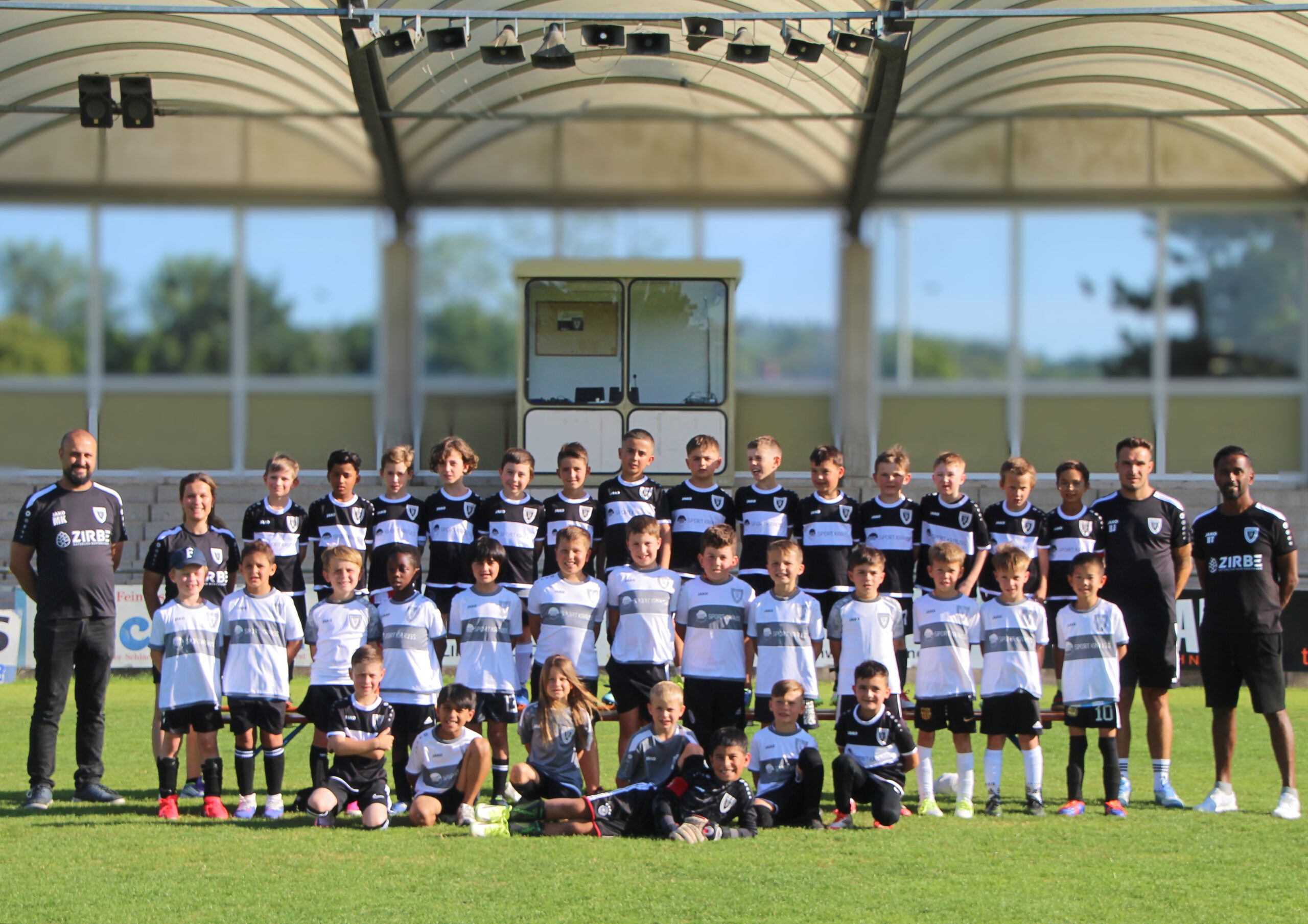 Group of soccer players and coaches in black-and-white uniforms posing for a team photo on a sunny field beneath a stadium structure.