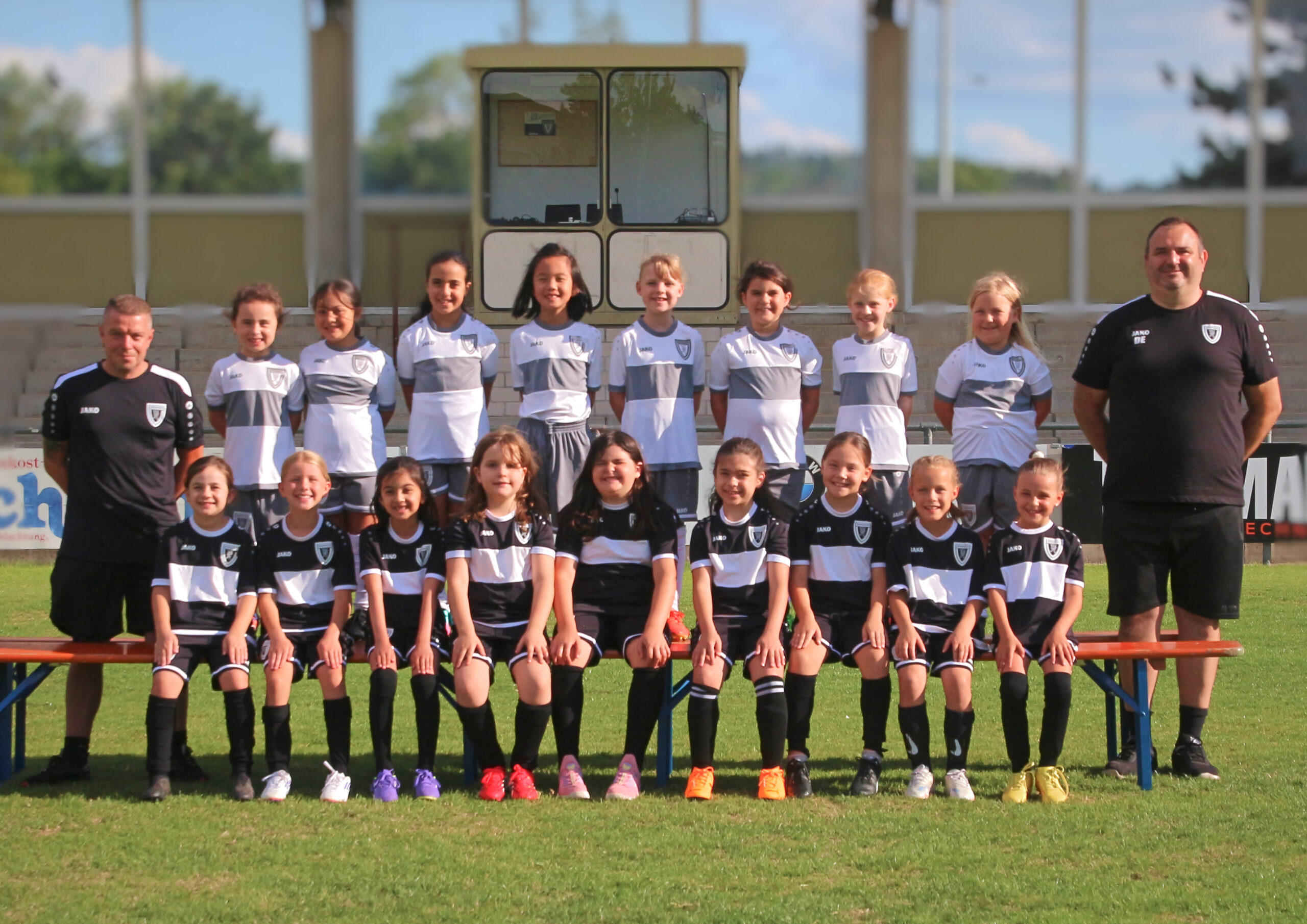 Youth soccer team posing for a group photo on a grassy field with two coaches on either end in black shirts and a line of players in white and gray uniforms behind them.